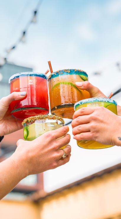 Four vibrant margarita-style cocktails with colorful sugared rims clinking in a rooftop toast on an urban patio with string lights and city buildings beyond