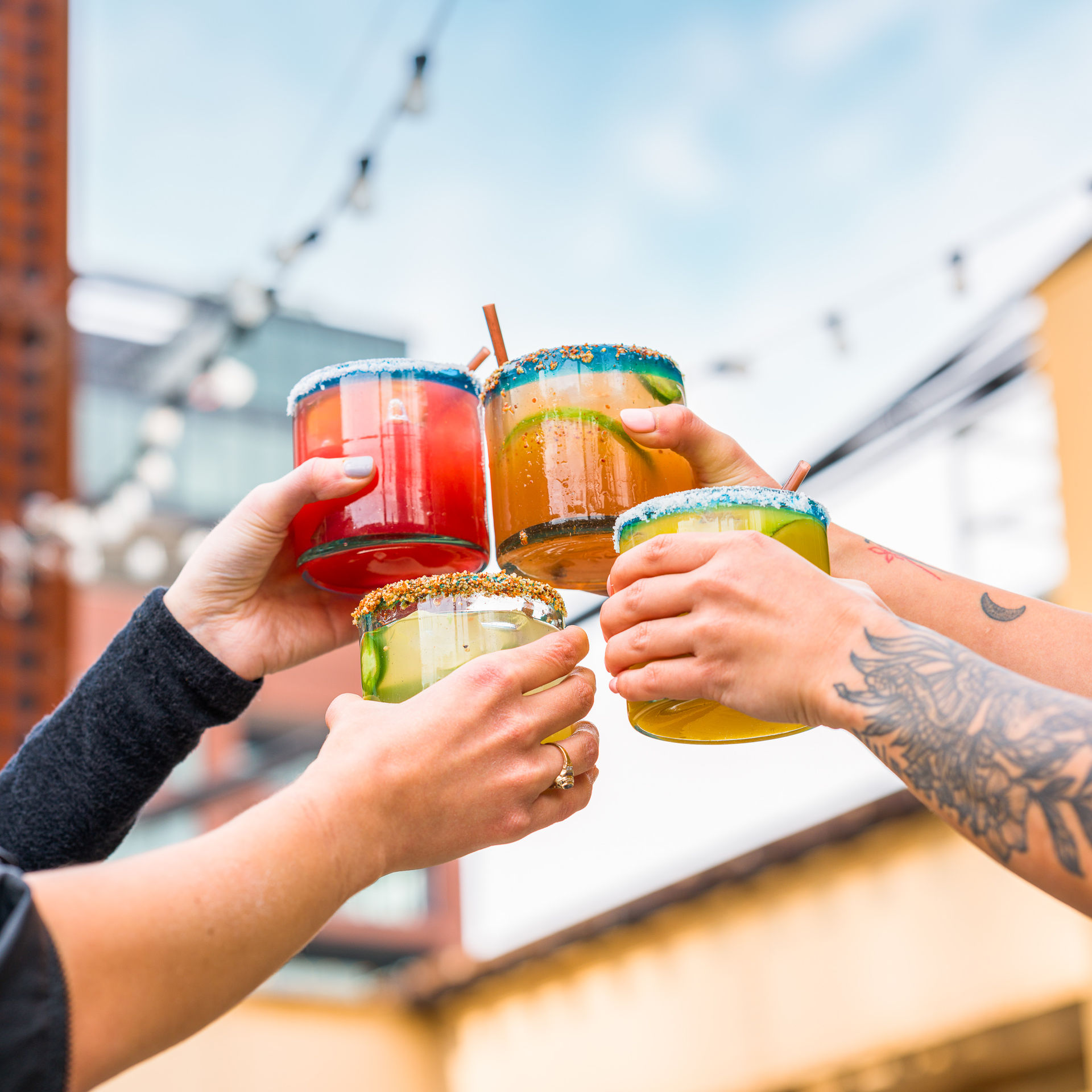 Four vibrant margarita-style cocktails with colorful sugared rims clinking in a rooftop toast on an urban patio with string lights and city buildings beyond