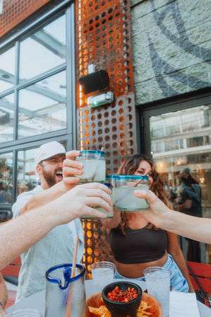 Friends clinking lime cocktails in blue-rimmed glasses over chips and salsa on an urban outdoor restaurant patio