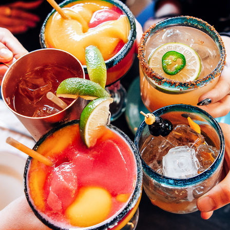 Vibrant cocktails clinking — frozen pink and mango margarita slushes with lime, a jalapeño‑topped margarita with chili salt rim, and an iced cocktail with a large ice cube.