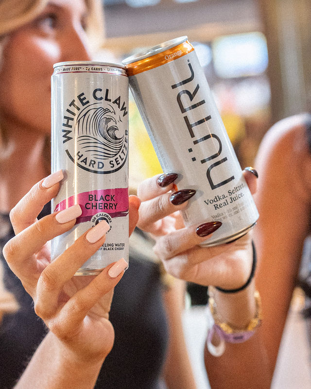 Close-up of two hard seltzer cans—black cherry and orange vodka seltzer—clinked together and held by hands with long manicured nails at a lively social gathering.