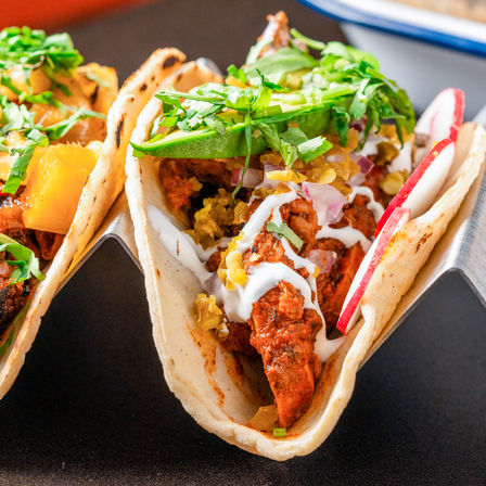 Close-up of street-style chicken tacos in soft flour tortillas topped with sliced avocado, pickled jalapeños, chopped red onion, radish, fresh cilantro and creamy white sauce.