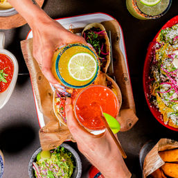 Top-down view of two hands clinking colorful margaritas—lime-garnished orange and red cocktails with salted rims—over a tray of street-style tacos, guacamole and nachos.
