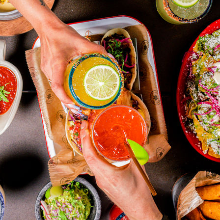 Top-down view of two hands clinking colorful margaritas—lime-garnished orange and red cocktails with salted rims—over a tray of street-style tacos, guacamole and nachos.