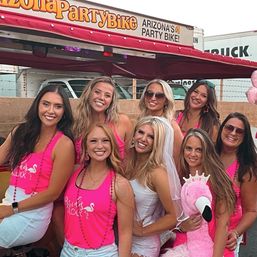 Eight women smiling on a bachelorette party bike in Arizona, most wearing matching hot-pink tank tops and bead necklaces; bride in white dress and veil holding a large pink flamingo plush.
