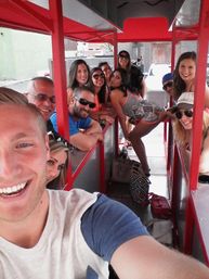 Smiling group taking a selfie aboard a red outdoor pedal party bike on a downtown street, friends laughing and posing under a canopy with bags on the floor.