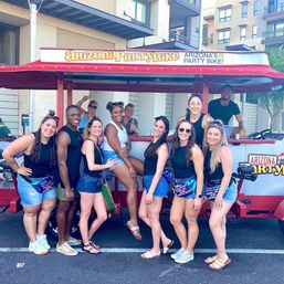 Cheerful group of friends in summer outfits posing beside a red pedal-powered party bike on a sunny downtown street with modern apartment buildings in the background.