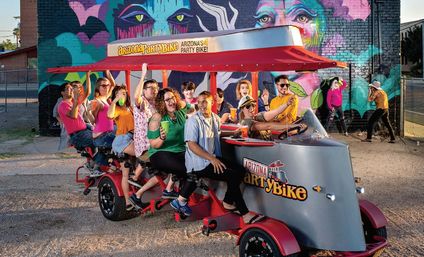 Group of friends pedaling a colorful multi-seat party bike on a downtown Arizona street, laughing and holding drinks in front of a vibrant mural of painted faces.