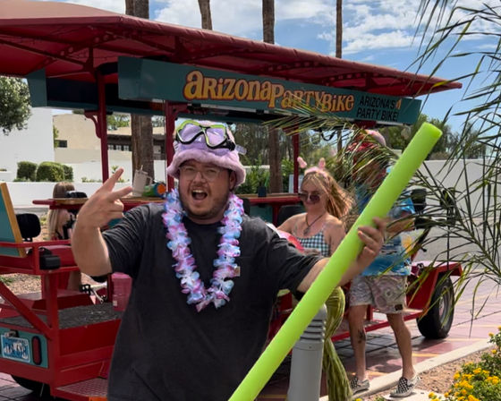 Person wearing a pink fuzzy hat with goggles and a floral lei, holding a bright green pool noodle and flashing a peace sign in front of a red party bike on a sunny Arizona street with palm trees.