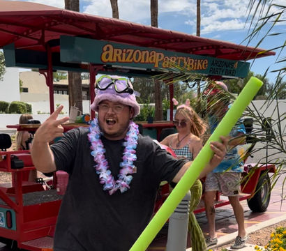 Person wearing a pink fuzzy hat with goggles and a floral lei, holding a bright green pool noodle and flashing a peace sign in front of a red party bike on a sunny Arizona street with palm trees.