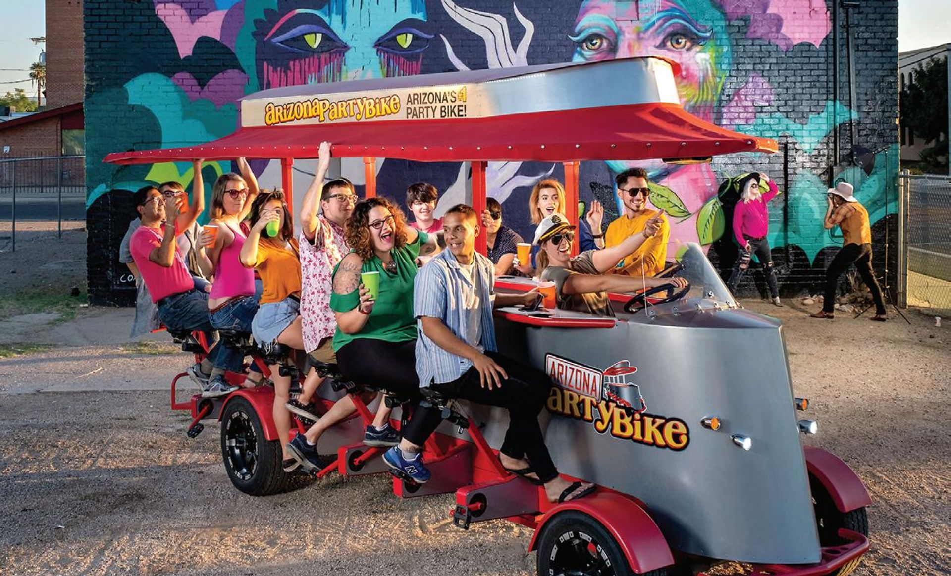 Laughing group of adults on a red pedal-powered party bike in front of a vibrant street-art mural, holding colorful cups and posing during an outdoor urban tour.