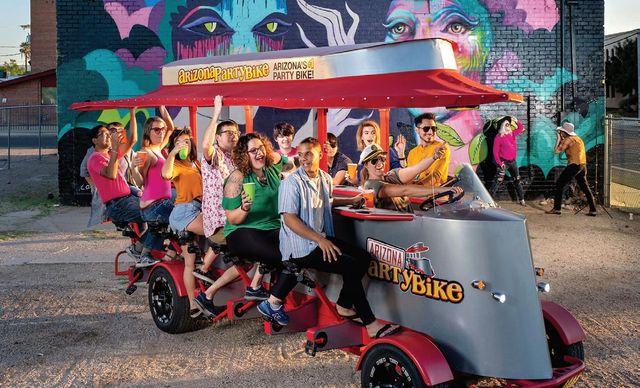 Laughing group of adults on a red pedal-powered party bike in front of a vibrant street-art mural, holding colorful cups and posing during an outdoor urban tour.
