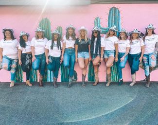 Group of women in cowgirl hats, denim and cowboy boots posing and smiling against a bright pink cactus mural for a fun Southwestern-style group photo.