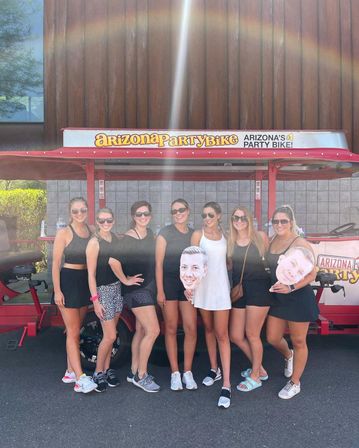 Eight women in summer outfits and sunglasses posing for a group photo in front of a red Arizona party bike with canopy, two holding oversized face cutouts