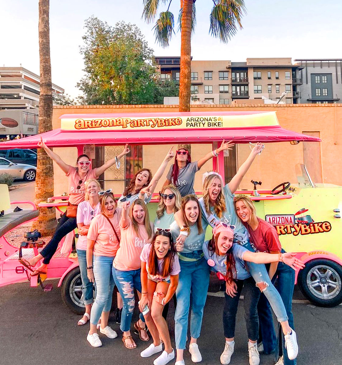 Bachelorette group of smiling women posing by a bright pink-and-yellow pedal party bike on a palm-lined Arizona street, celebrating outdoors