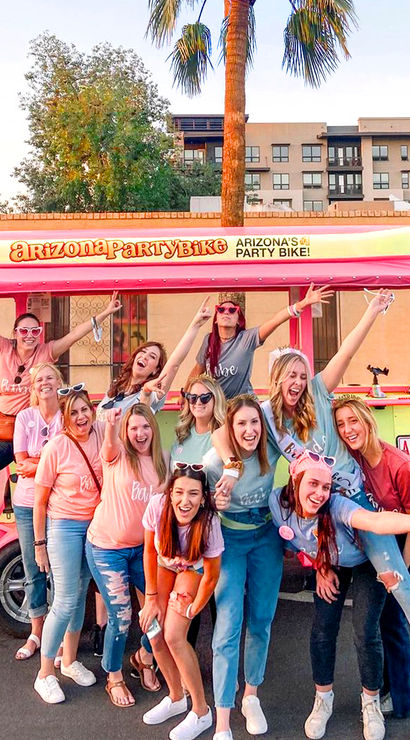 Smiling group of women in colorful shirts posing for a bachelorette-style outing in front of a pink party bike on a sunny Arizona street with palm trees and apartments