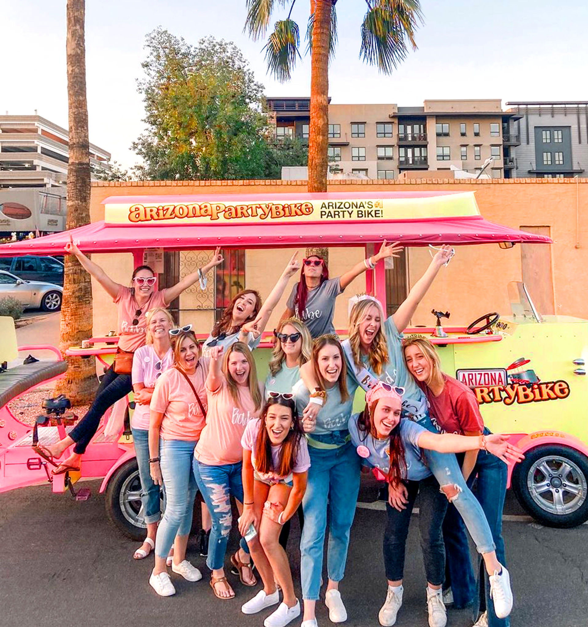 Smiling group of women in colorful shirts posing for a bachelorette-style outing in front of a pink party bike on a sunny Arizona street with palm trees and apartments