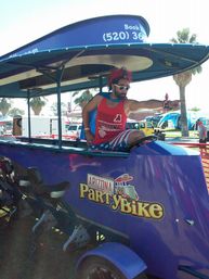 Person in red tank top, star-spangled shorts and white sunglasses striking a pose and pointing from a purple pedal-powered party bike at an outdoor Arizona festival with palm trees