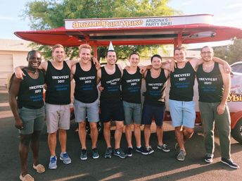 Eight friends in matching “Trevfest 2017” tank tops smiling and posing in front of a red open-air pedal party bike on a sunny Arizona street.