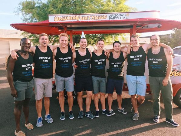 Eight friends in matching 'TREVFEST 2017' tank tops smiling and arm-in-arm in front of a red party-bike vehicle in a sunny parking lot, one wearing a cone party hat.