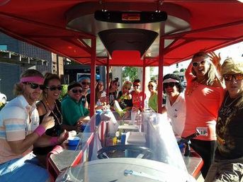 Cheerful group of a dozen people wearing sunglasses and colorful outfits riding a red party bike/pedal pub with drinks under a canopy on a sunny downtown street.
