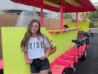 Smiling woman wearing a 'BRIDE' T-shirt posed beside a neon yellow and hot-pink pedal party bike with multiple seats in an apartment parking lot.