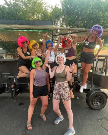 Seven women wearing colorful wigs and sunglasses laughing and posing on a parked party bike in a tree-lined outdoor parking area — playful summer group photo