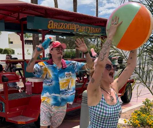 Two adults celebrating on a colorful open-air party bike in sunny Arizona — woman in a checkered dress tosses a giant beach ball while a man in a tropical shirt and pink cap fires a water gun.