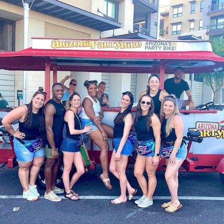 Smiling group of adults posing in front of a red pedal-powered party bike on a sunny Arizona street with modern apartment buildings in the background.