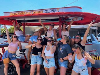 Group of young women in summer outfits drinking, posing, and flashing peace signs on a red outdoor pedal-powered party bike in sunny Arizona