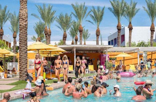 Sunny resort pool party with tall palm trees and yellow umbrellas; crowded outdoor pool with people in swimwear lounging by cabanas and socializing in the water — lively vacation scene.