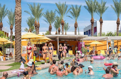 Sunny resort pool party with tall palm trees and yellow umbrellas; crowded outdoor pool with people in swimwear lounging by cabanas and socializing in the water — lively vacation scene.
