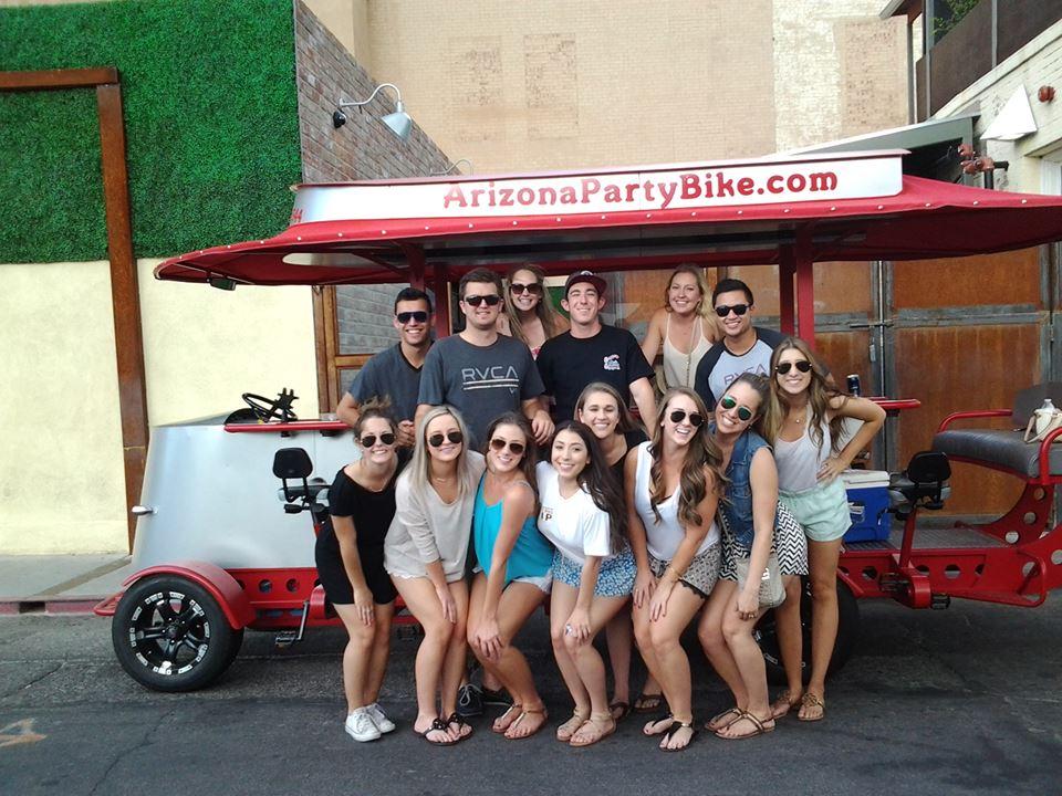Cheerful group of young adults posing in front of a red pedal-powered party bike on a sunny urban street, wearing summer outfits and sunglasses.