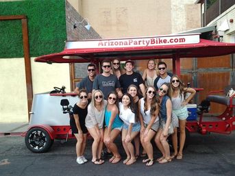 Happy group of friends posing by a red pedal-powered party bike on a sunny Arizona street, ready for a city pub crawl.