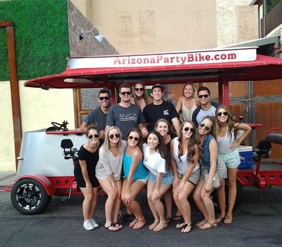Cheerful group of young adults posing in front of a red pedal-powered party bike on a sunny urban street, wearing summer outfits and sunglasses.