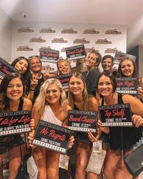 Smiling group of women at a girls' night bachelorette photo booth, posing with mugshot-style novelty signs and playful captions against a party backdrop.