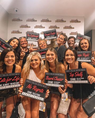 Smiling group of women at a girls' night bachelorette photo booth, posing with mugshot-style novelty signs and playful captions against a party backdrop.