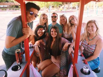 Eight young adults smiling on a red outdoor party bike (pedal pub) on a sunny day, wearing sunglasses and summer outfits with canned drinks in cup holders