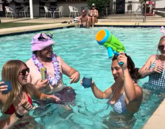 Friends enjoying a sunny outdoor pool party in swimsuits with drinks, a lei, sunglasses, and a colorful water gun.