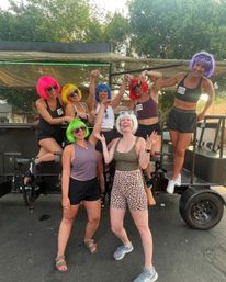 Seven women wearing bright neon wigs and sunglasses laughing and posing on a pedal‑pub party bike parked on a sunny street — playful summer group outing