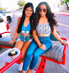Two friends smiling on a red outdoor bench on a sunny city street, wearing summer crop tops and denim (jeans and shorts), sunglasses, with a soda can in the cupholder