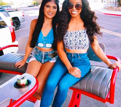 Two smiling friends in summer crop tops and denim (shorts and high-waisted jeans) sitting on a red outdoor bench by a sunny downtown street, sunglasses and a soda can in a cup holder.