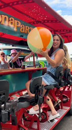 Smiling woman on a red outdoor pedal bar holding a colorful beach ball under a festive canopy during a summer group ride