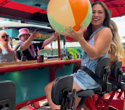 Smiling woman on a red outdoor pedal bar holding a colorful beach ball under a festive canopy during a summer group ride