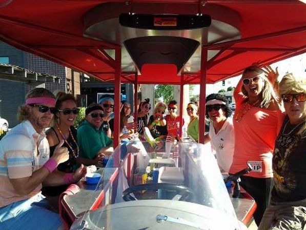 Group of smiling people on a red pedal-powered party bike (pedal pub) under a canopy, wearing sunglasses and bright outfits, drinking and socializing on a sunny urban street.