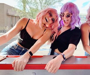 Two friends rocking pastel pink and purple wigs and cat-eye sunglasses, smiling and leaning on a red railing in a sunny outdoor setting, wearing black tops and denim.