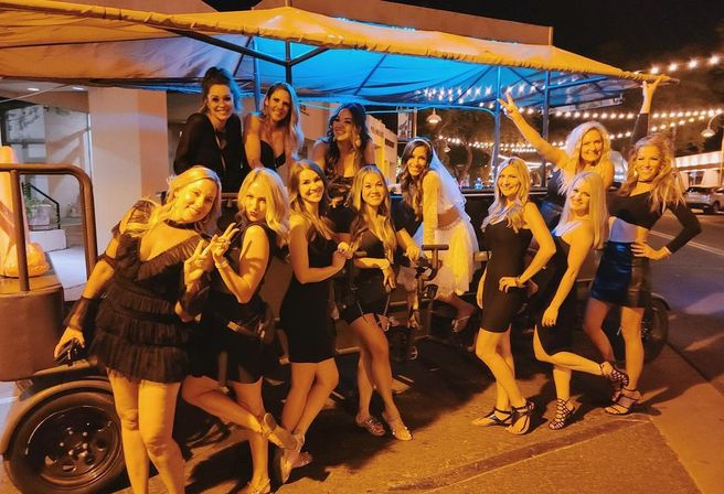 Group of women, mostly in black dresses and one in a white bridal dress, posing on a covered party bike/pedal pub under string lights on a downtown street at night