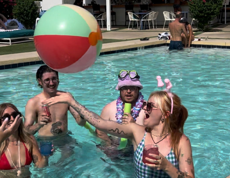 Sunny outdoor swimming pool party with young adults in swimsuits tossing a large colorful beach ball; one person wears a pink novelty headband and sunglasses, another sports goggles and a lei, guests laughing and holding colorful drinks.