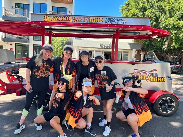Seven friends in matching flame-print shirts pose and point in front of a red pedal-powered party bike on a sunny Arizona street.