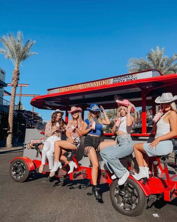 Five women in cowboy hats and boots riding a red pedal party bike on a sunny Arizona street lined with palm trees, sipping drinks and posing for a fun group outing.